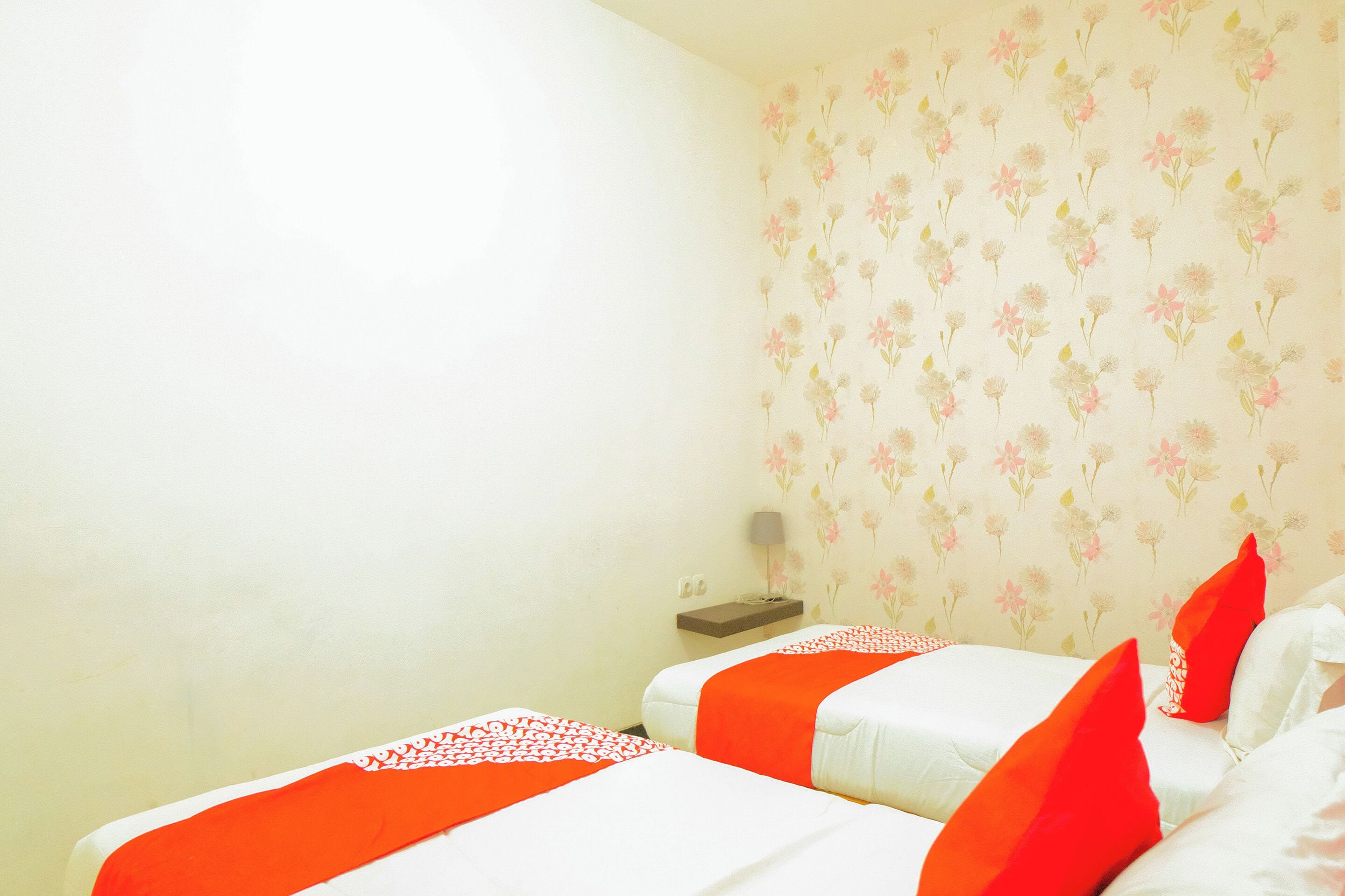 A hotel room with two beds, each with white bedding and orange accents. The room has a floral wallpaper and a small bedside table with a lamp.