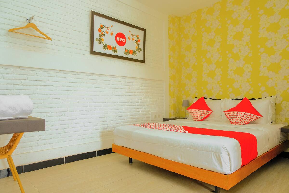 A hotel room with a white brick wall, a framed picture with a floral wreath, a bed with white and red bedding, and a yellow floral wallpaper.