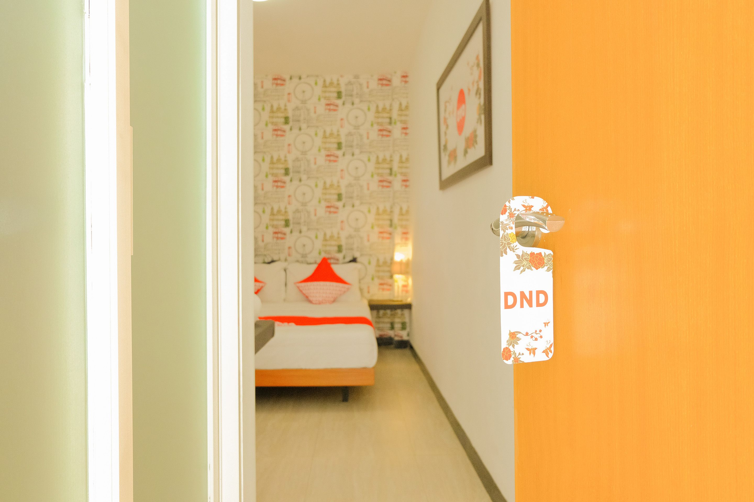 Un pasillo estrecho con una cama al fondo, una puerta con el letrero 'DND' y un papel tapiz estampado.