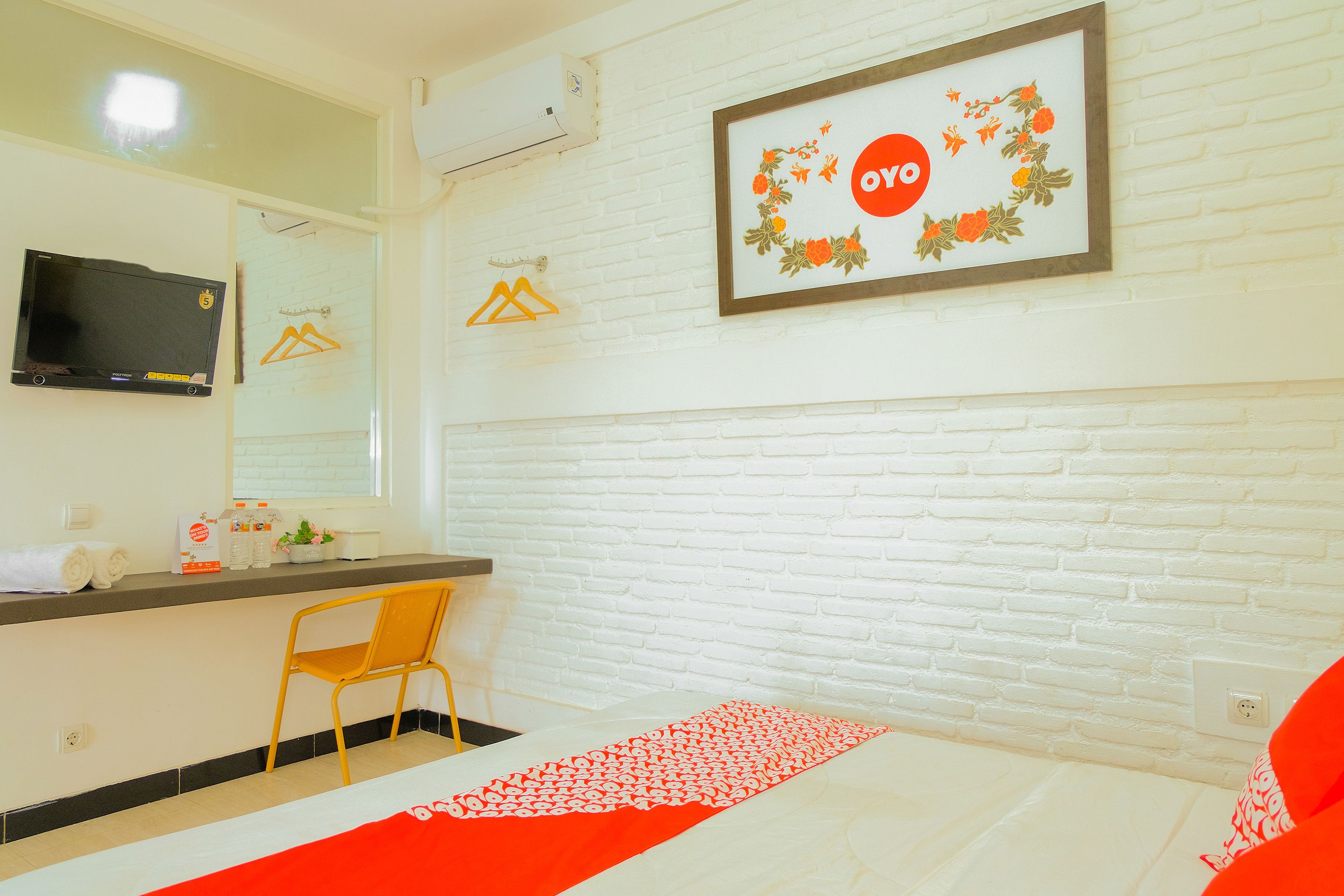 A hotel room with a white brick wall, a bed with a red and white bedspread, a wooden chair, a TV mounted on the wall, and a framed picture with a floral design.
