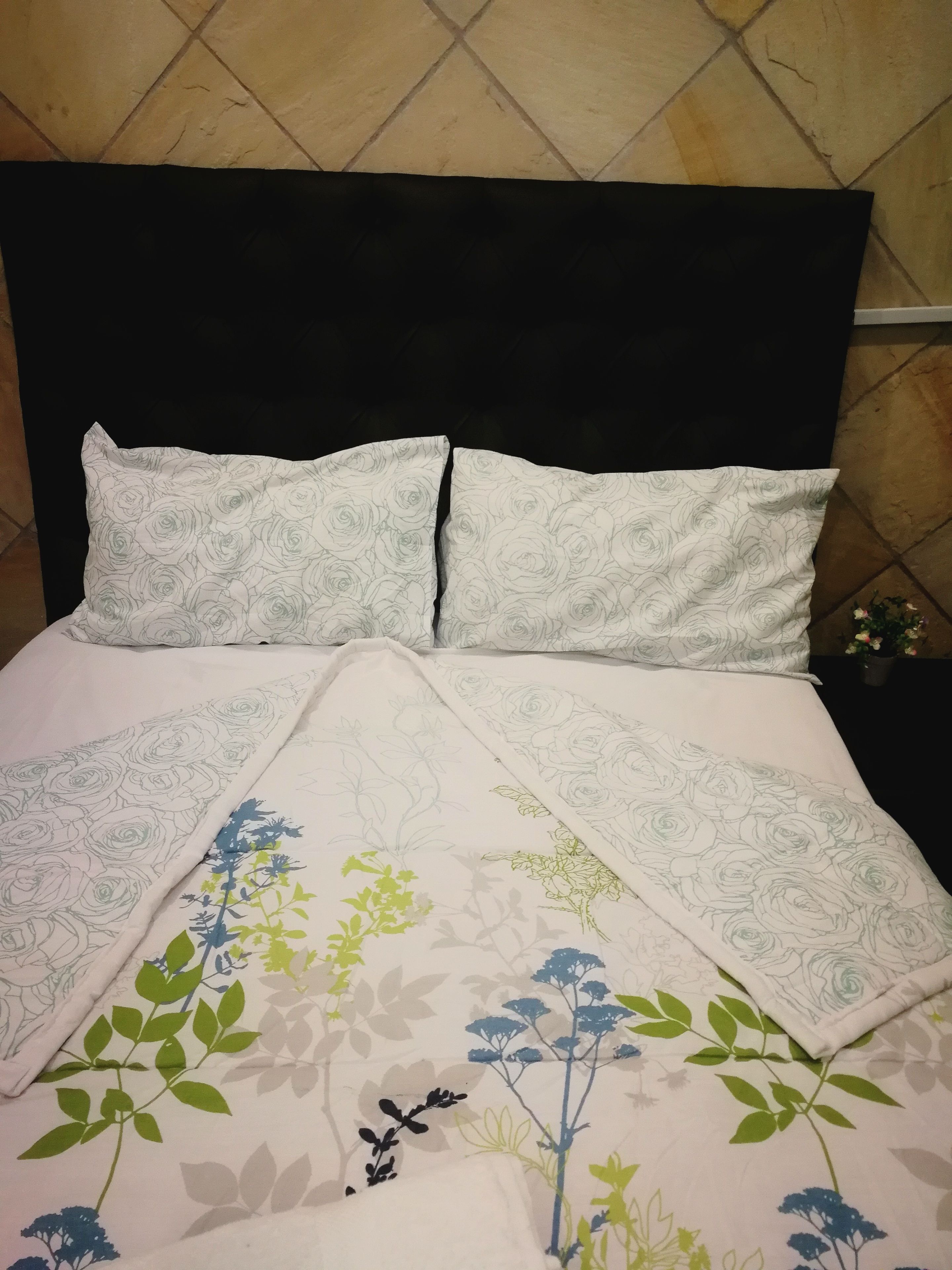 A bed with a black headboard, white pillows with a rose pattern, and a floral bedspread with green and blue flowers.