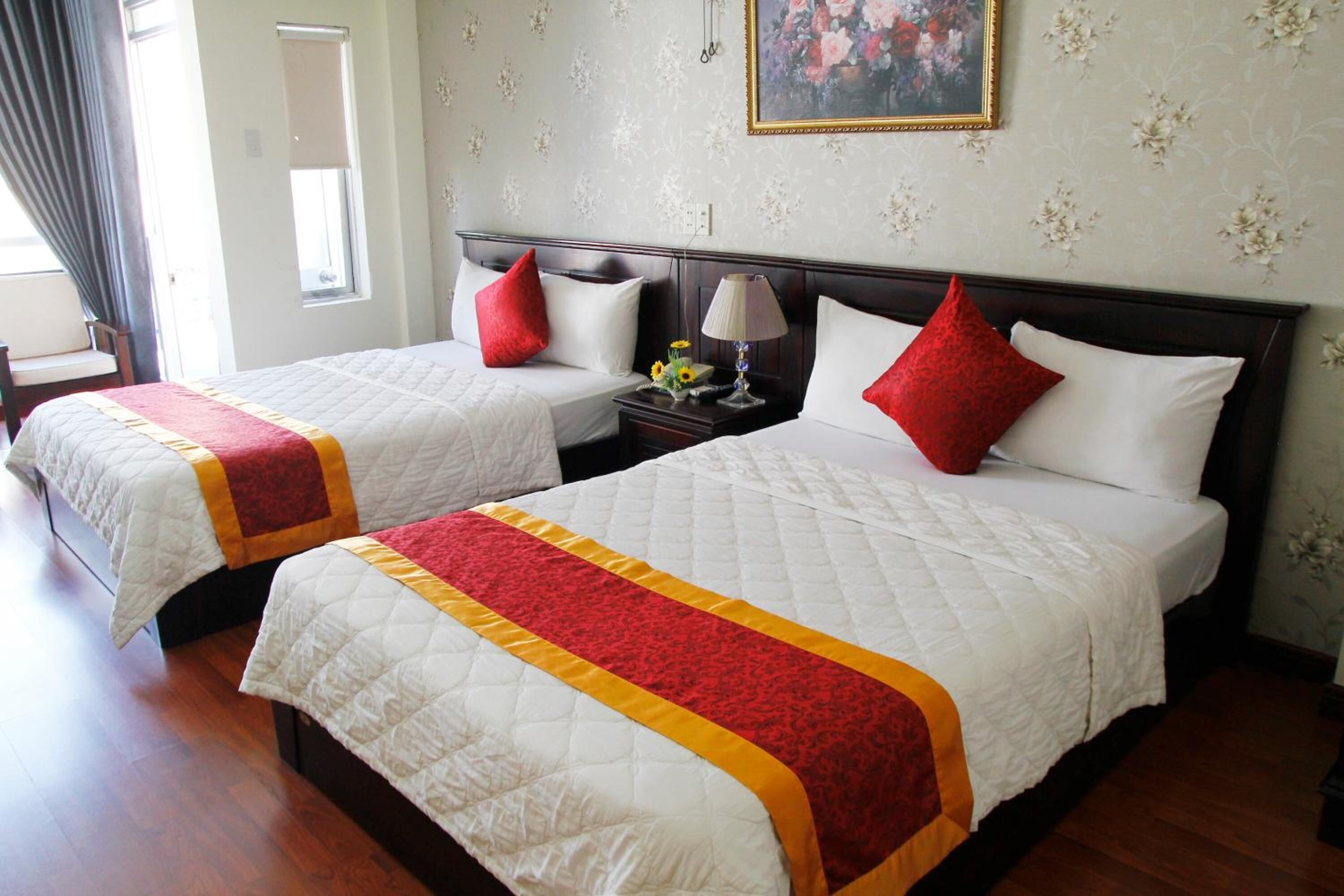 A hotel room with two beds, each with a white quilt and red accents, a nightstand with a lamp, and a floral wallpaper.