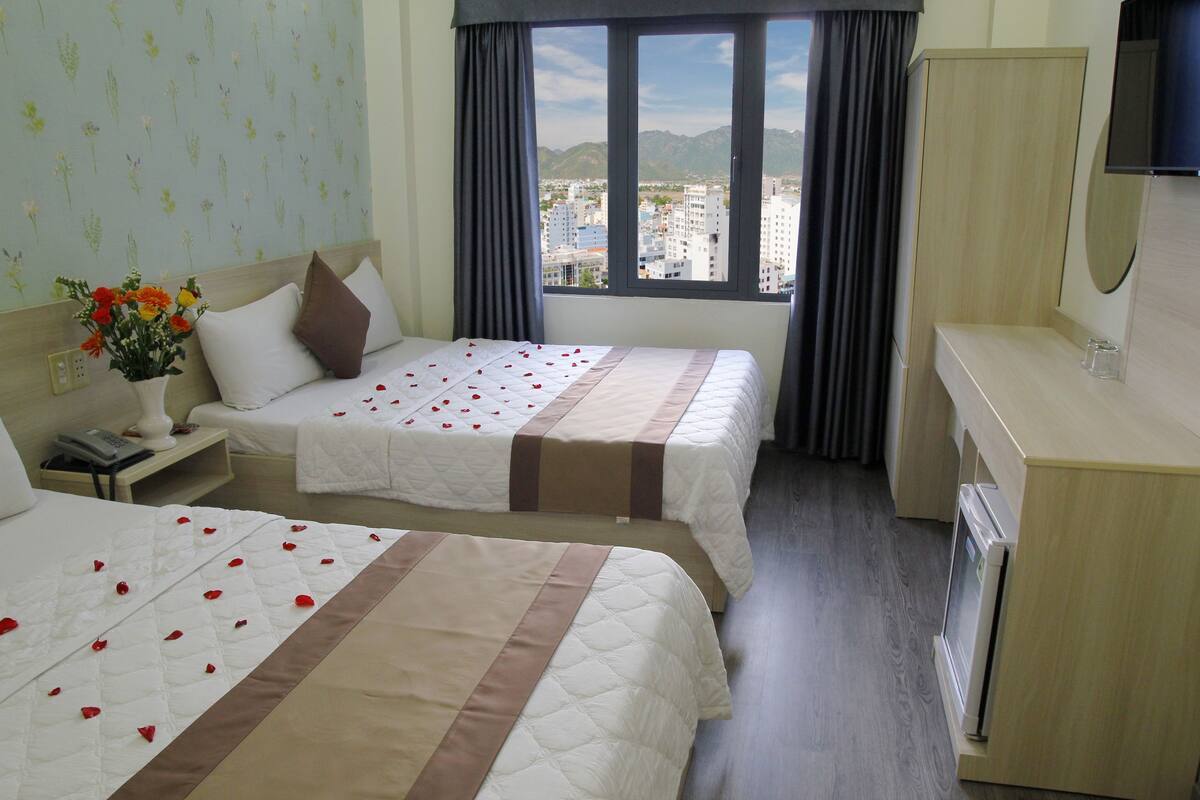 Een hotelkamer met twee bedden, een nachtkastje met een lamp, een klein tafeltje met een bloevas, een groot raam met uitzicht op gebouwen, en een magnetron.