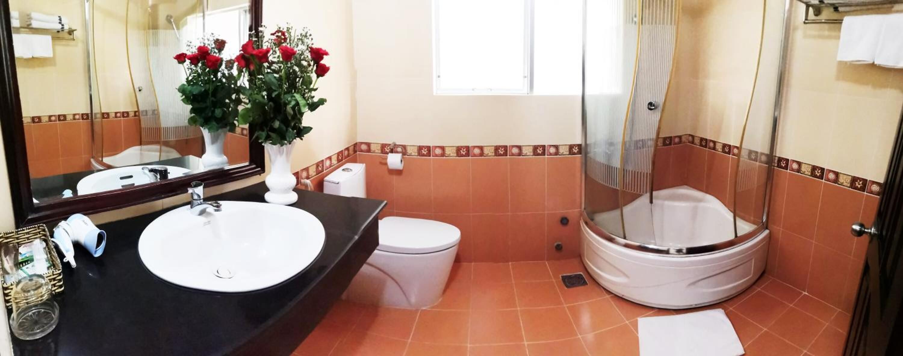 A bathroom with a round sink, toilet, and bathtub. There is a mirror, a vase with flowers, and a shower curtain.