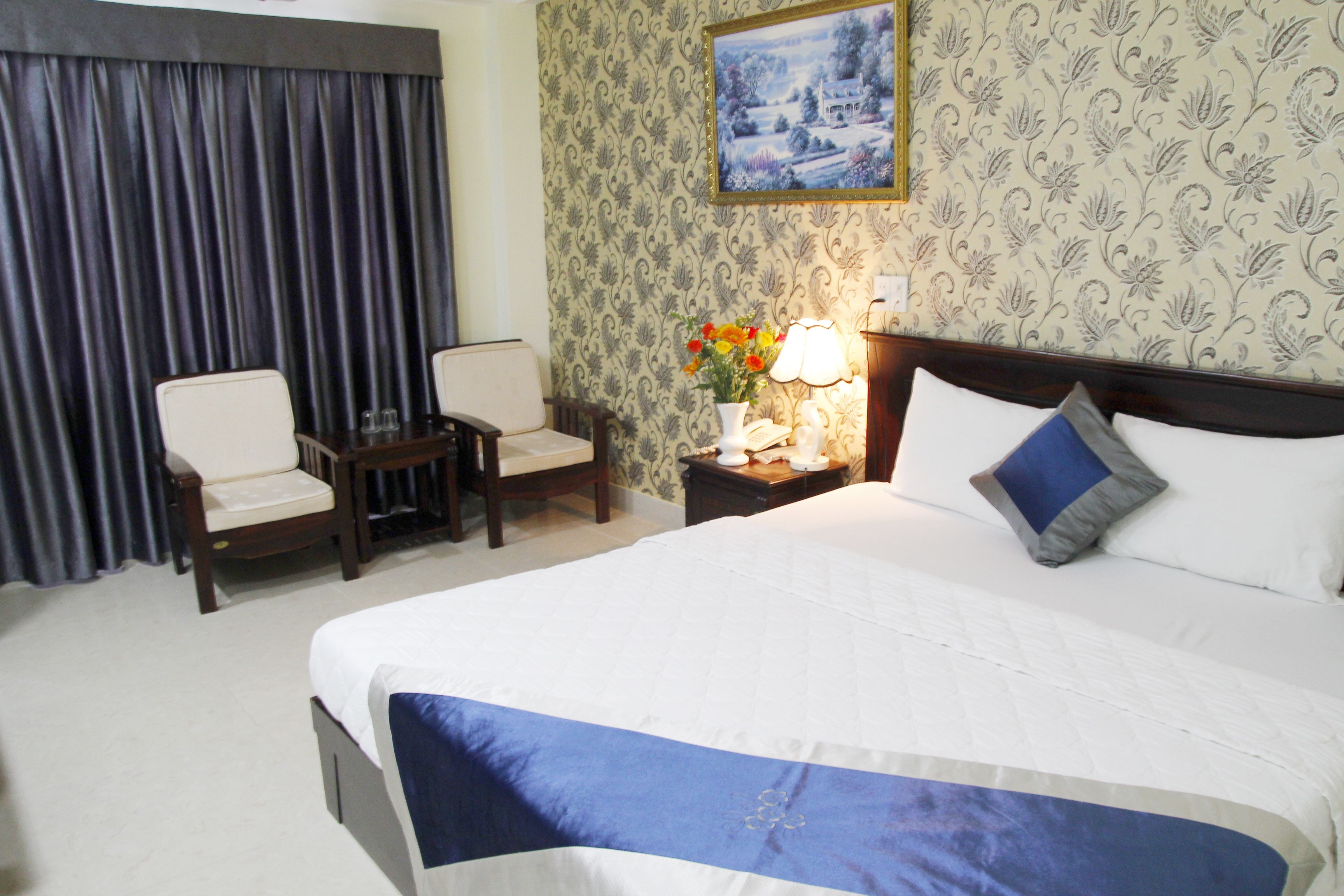 A hotel room with a large bed, two chairs, a nightstand with a lamp, and a painting on the wall.