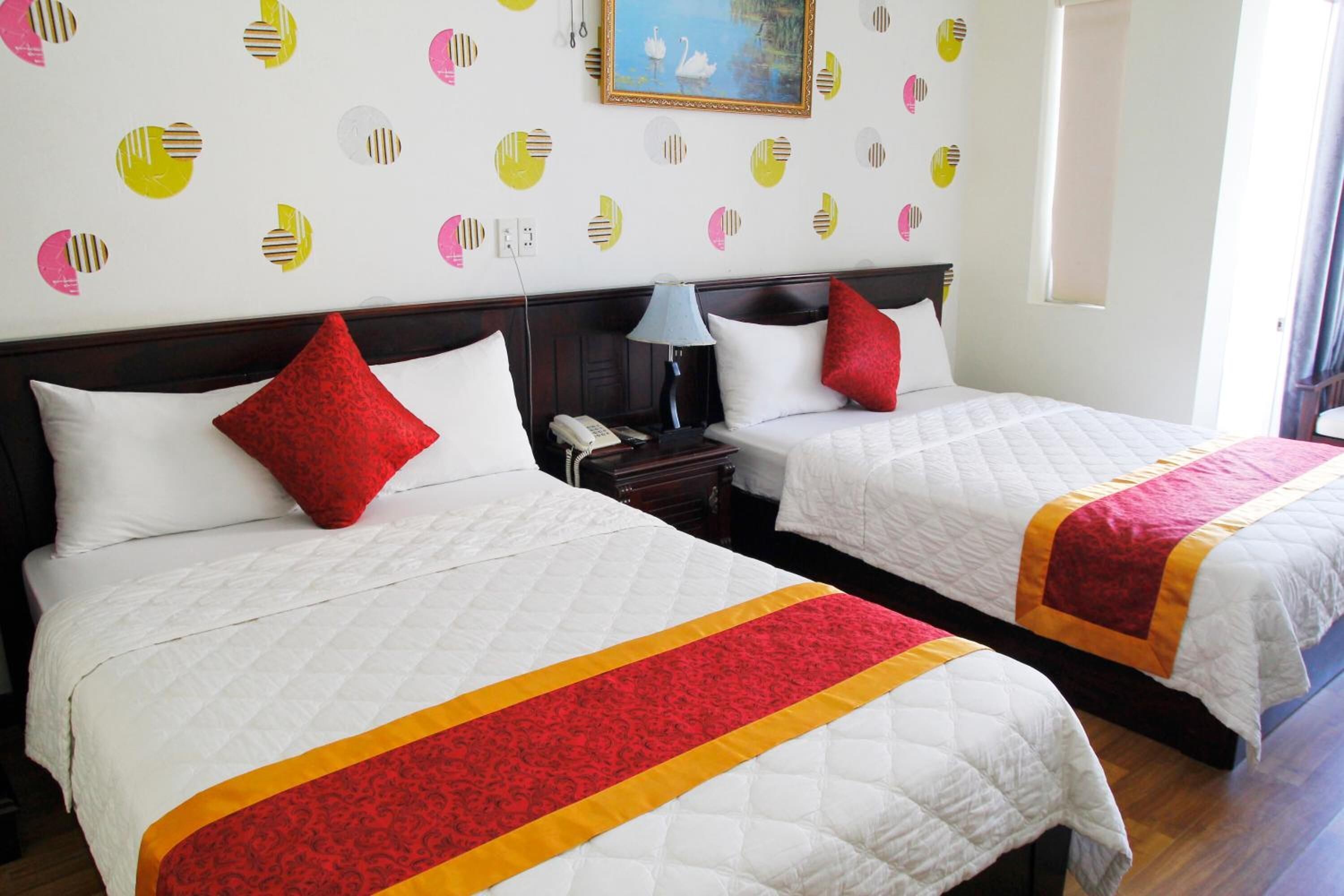 Two beds with white bedding and red accents, a nightstand with a lamp, and a wall with circular patterns.