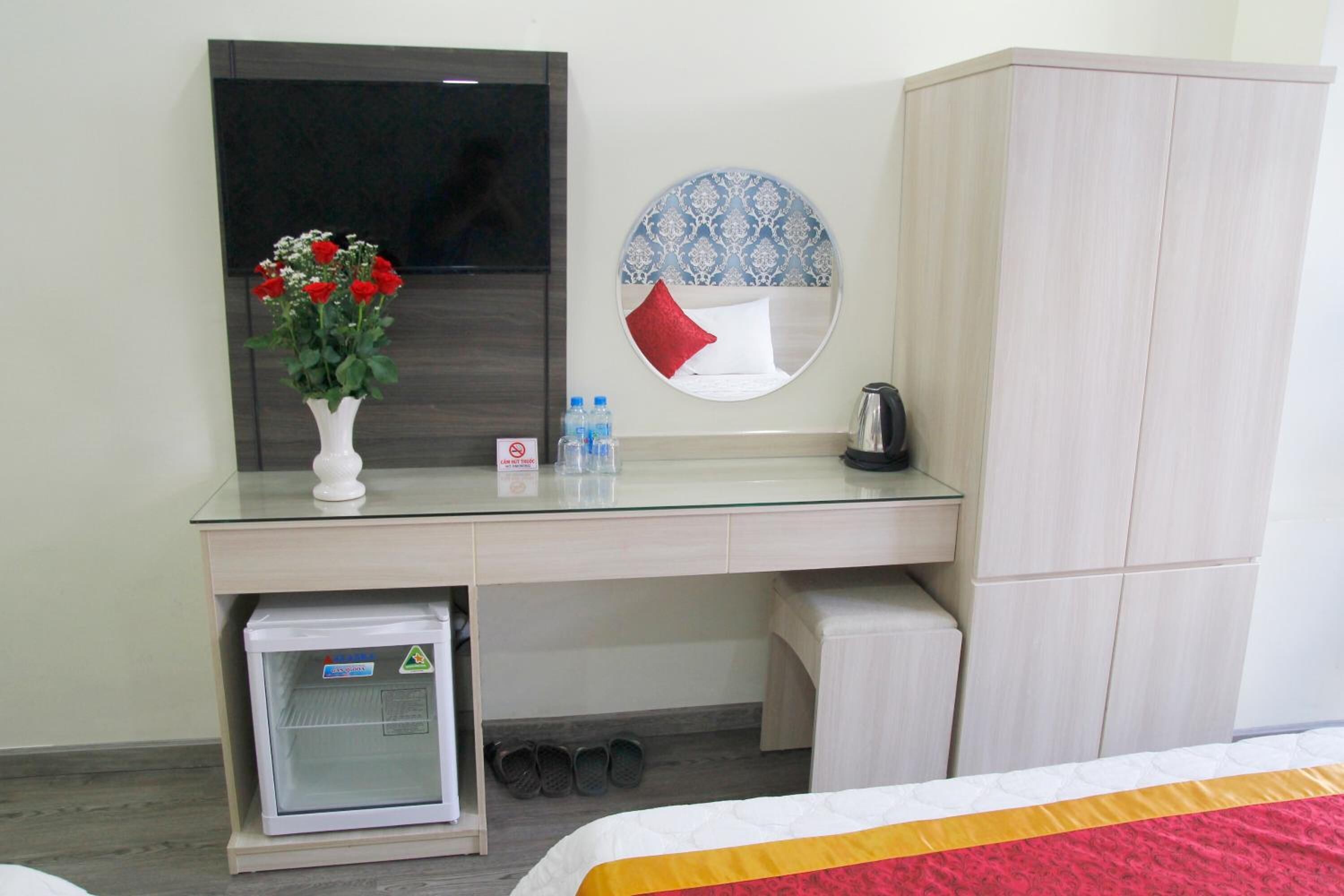 A hotel room with a glass desk, a small refrigerator, a bed with a red and yellow bedspread, a wardrobe, a round mirror with a blue and white pattern, and a vase with red flowers.