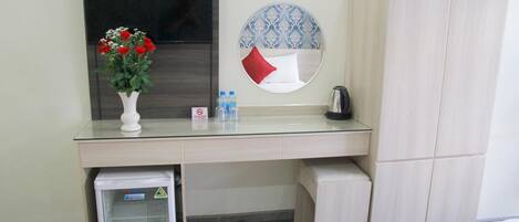 A hotel room with a glass desk, a small refrigerator, a bed with a red and yellow bedspread, a wardrobe, a round mirror with a blue and white pattern, and a vase with red flowers.