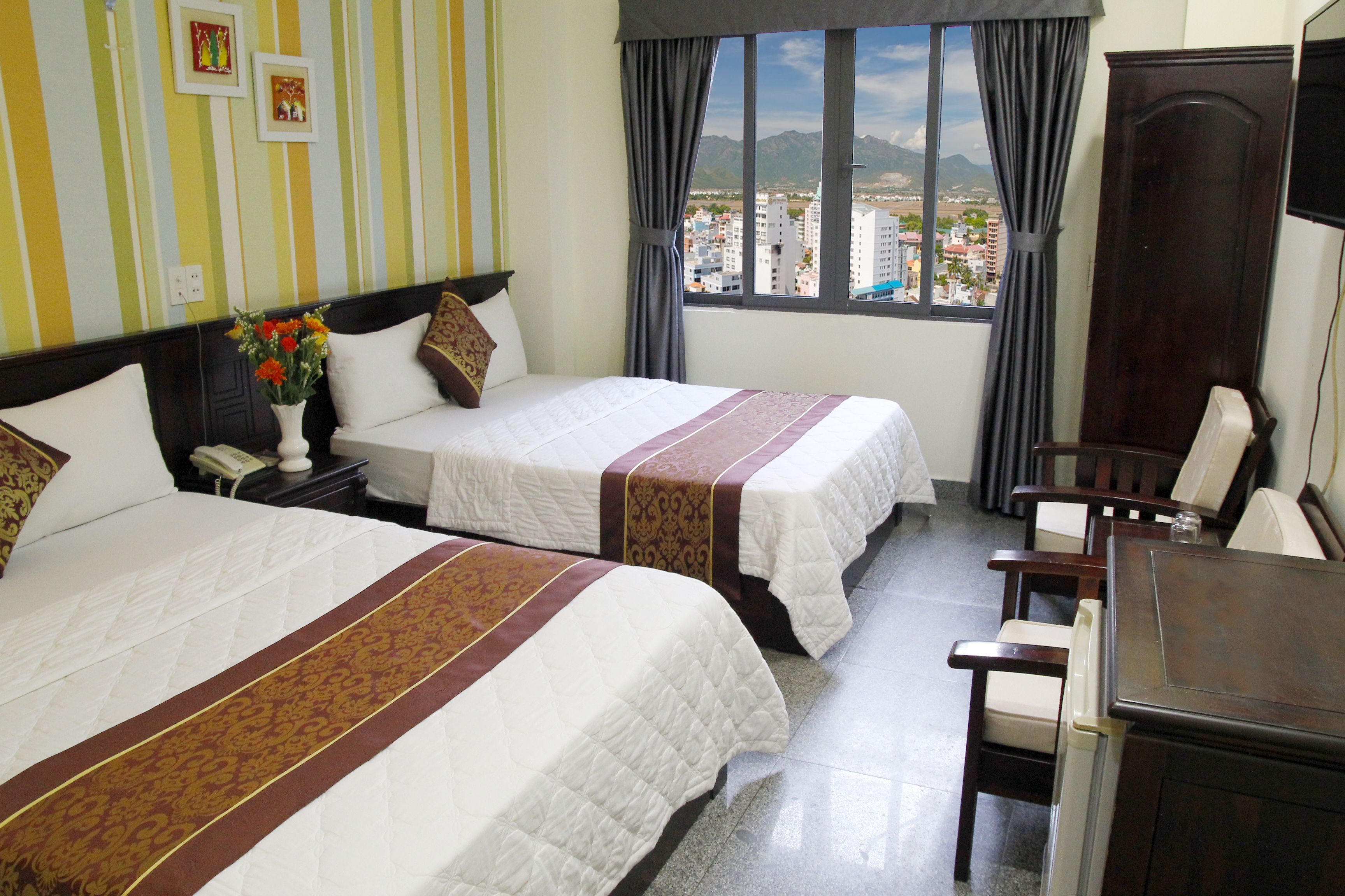 A hotel room with two beds, a desk, and a chair. There is a window with a view of buildings and mountains.