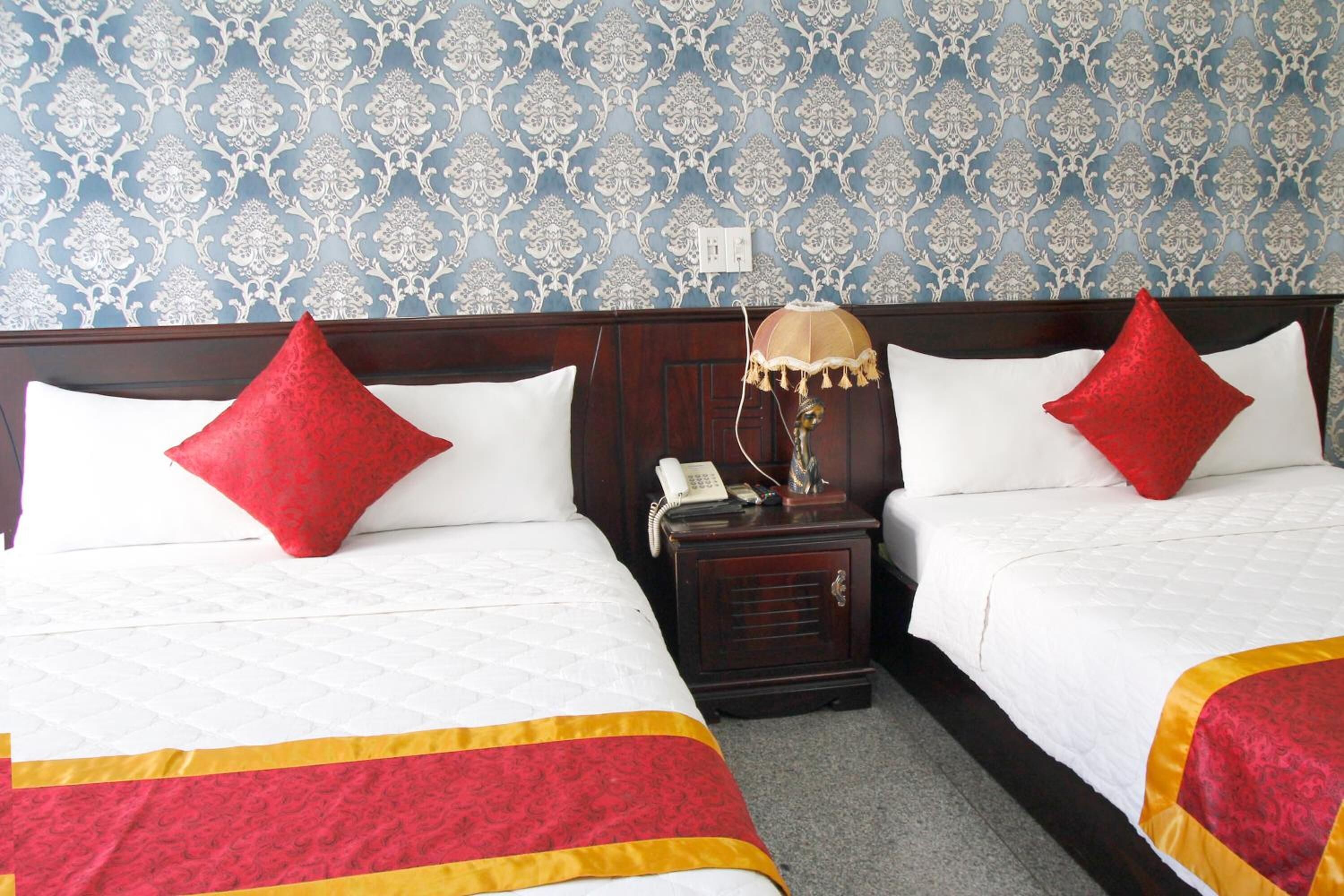 A hotel room with two beds, each with a red pillow and a white bedspread. There is a wooden nightstand with a lamp and a telephone between the beds. The wall has a blue patterned wallpaper.