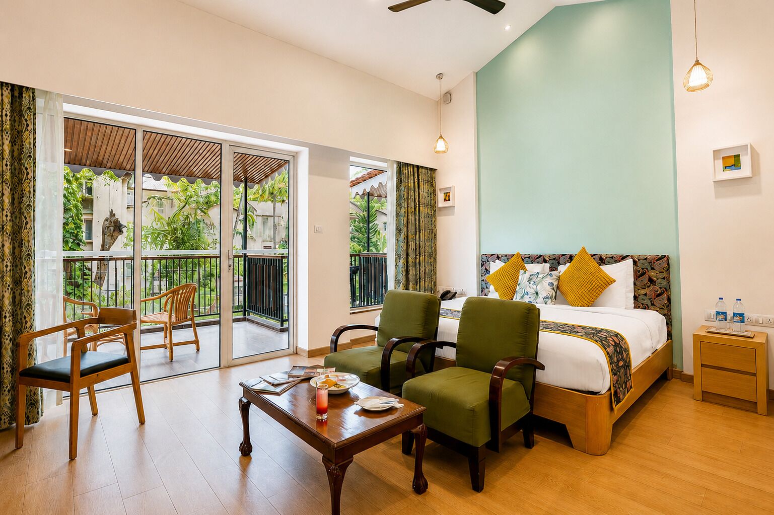 Habitación de hotel con cama, dos sillones, una mesa de madera, una mesita de noche y un balcón con vistas a zonas verdes.