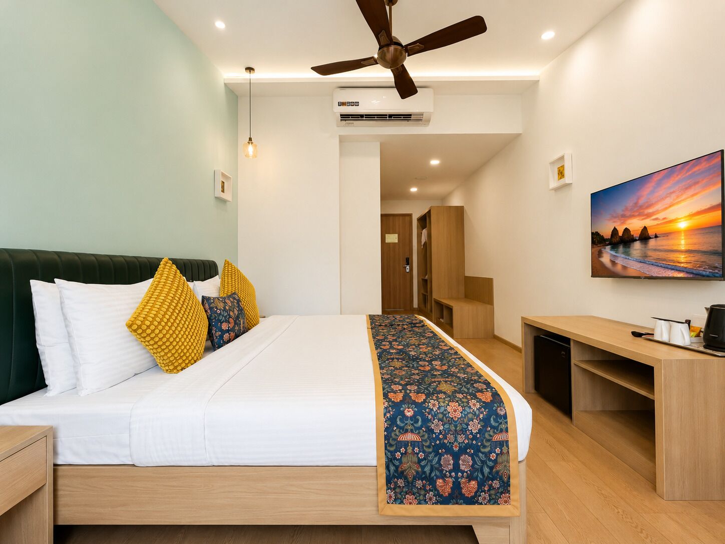 Habitación de hotel con cama, escritorio, ventilador de techo, aire acondicionado, un cuadro y una puerta que da a otra habitación.