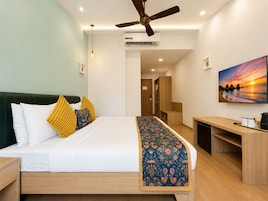 Habitación de hotel con cama, escritorio, ventilador de techo, aire acondicionado, un cuadro y una puerta que da a otra habitación.