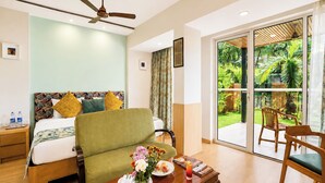 A hotel room with a bed, a green armchair, a dining table with a vase of flowers, and a view of a garden through a sliding glass door. - The BeachWalk Resort by Acron (Candolim)