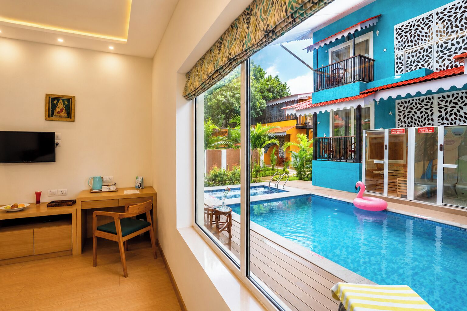 Habitación con una ventana grande que da a una piscina, un escritorio de madera, un televisor y una mesita con una taza y un plato.