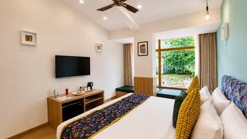 Habitación de hotel con cama, escritorio con televisor de pantalla plana, ventilador de techo y ventana con vistas a la vegetación.