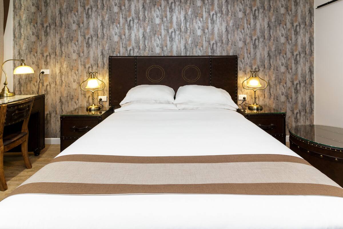A neatly made bed with a brown and white striped bedspread, two bedside lamps, a wooden chair, and a small table with a lamp.