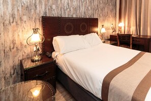 Habitación de hotel con cabecera de madera, cama con ropa blanca, dos lámparas de noche, mesita de noche de madera y una mesa de vidrio con lámpara.