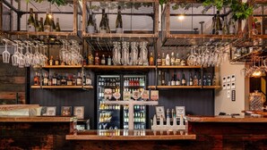Zona de bar con estantes llenos de botellas de licor, copas colgando y un refrigerador con bebidas.