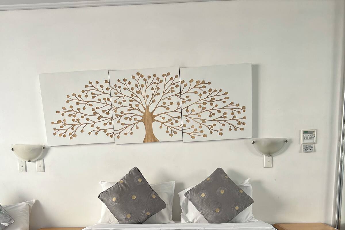 Una cama bien hecha con sábanas blancas y dos almohadas, una lámpara de pared y un cuadro de un árbol.
