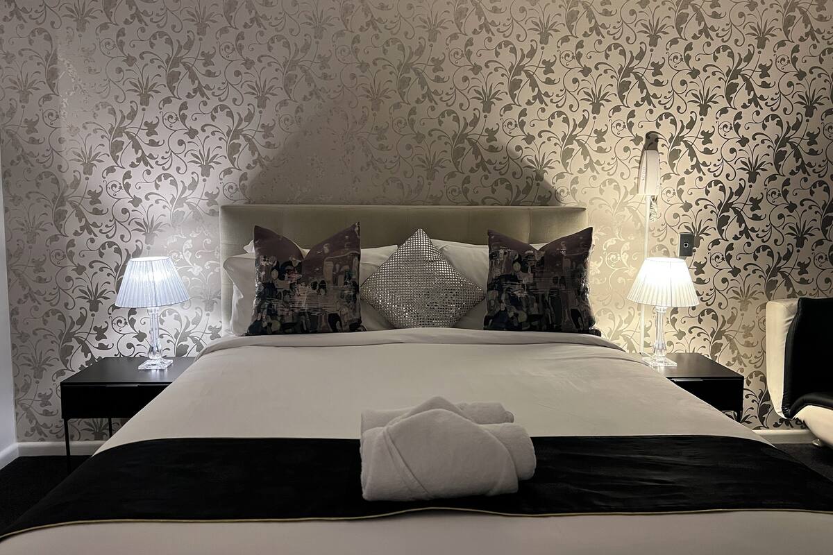Una cama bien hecha con edredón blanco y almohadones decorativos, una mesita de noche con lámpara, y una pared con estampado floral.