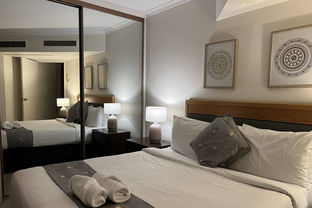 Habitación de hotel con una cama grande, dos mesitas de noche con lámparas, un espejo y una obra de arte en la pared.