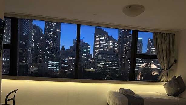 A hotel room with a large window offering a cityscape view at dusk. - Sydney Hyde Park Paxsafe Apartments (Darlinghurst)