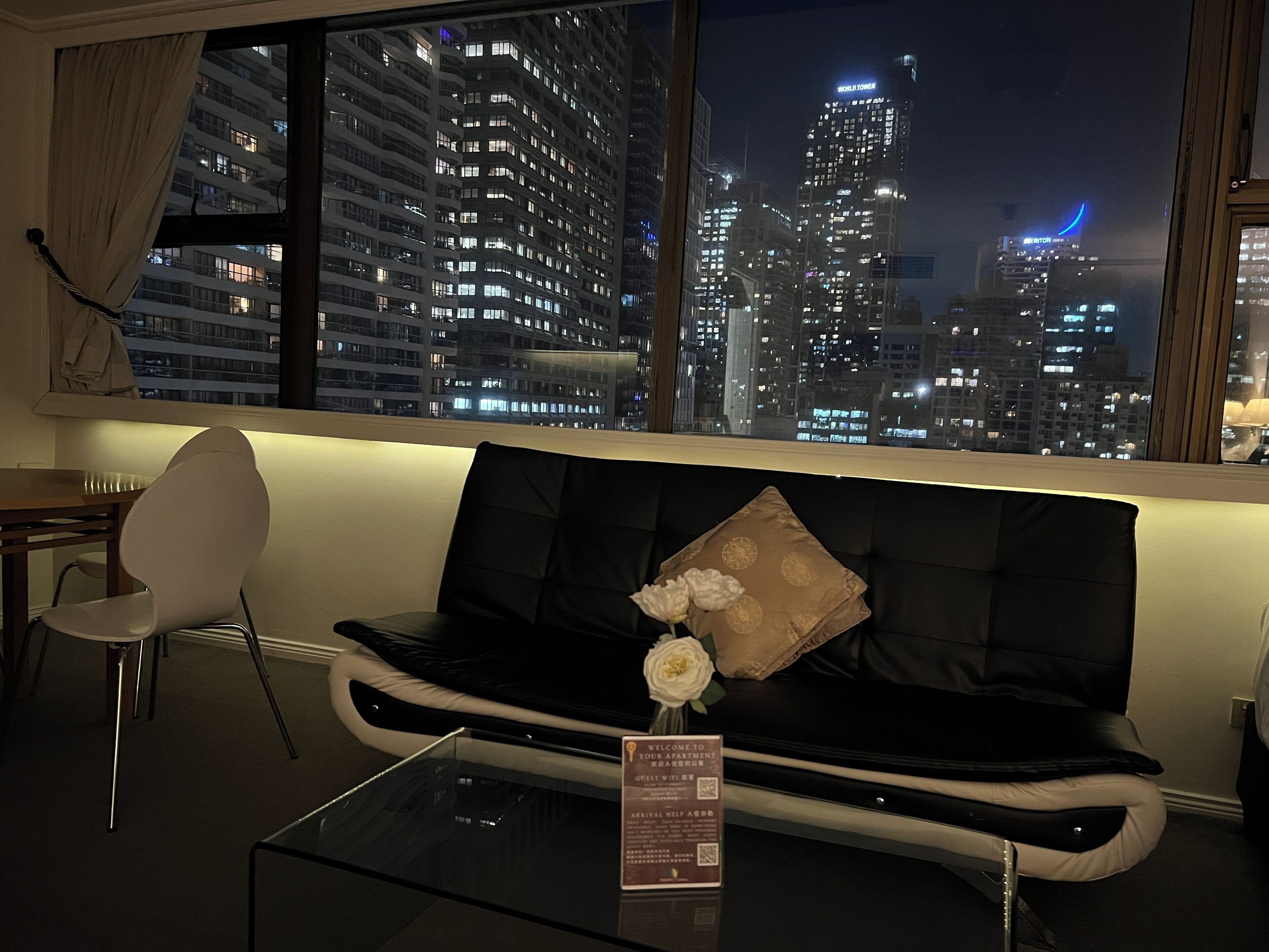 Un soggiorno moderno con un divano nero, un tavolino in vetro e vista sullo skyline cittadino di notte.
