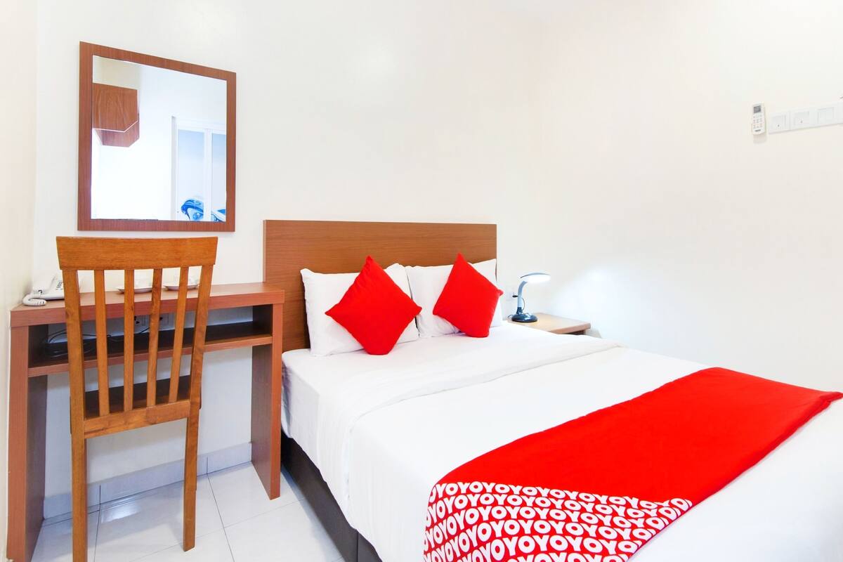 Una habitación de hotel con una cama, un escritorio, una silla, un espejo y una mesita de noche con una lámpara.