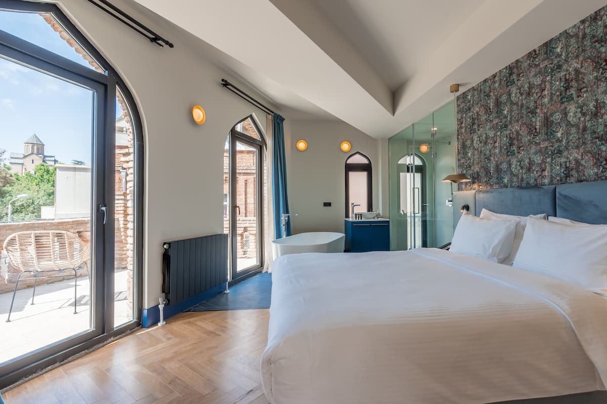 A modern bedroom with a large bed, a bathroom with a freestanding tub, and a view of a cityscape through large windows.