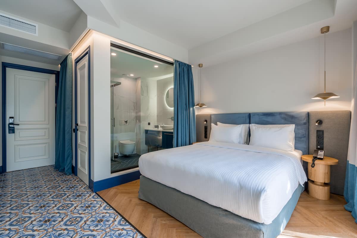 A modern hotel room with a large bed, a walk-in closet, a bathroom with a glass shower, and a small bedside table with a lamp.