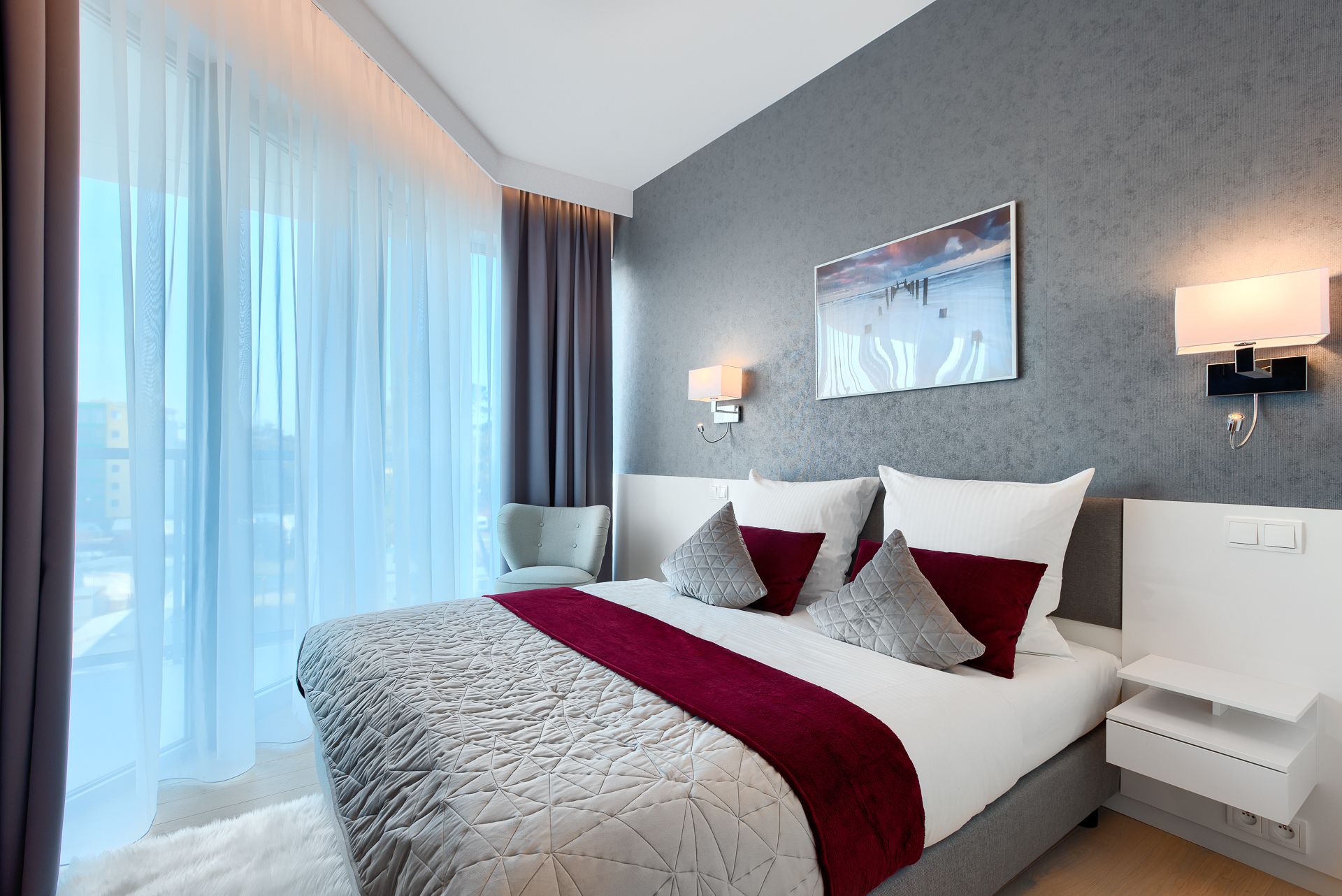 A modern hotel room with a large bed, a view of the city through sheer curtains, a grey textured wall, and a painting above the bed.