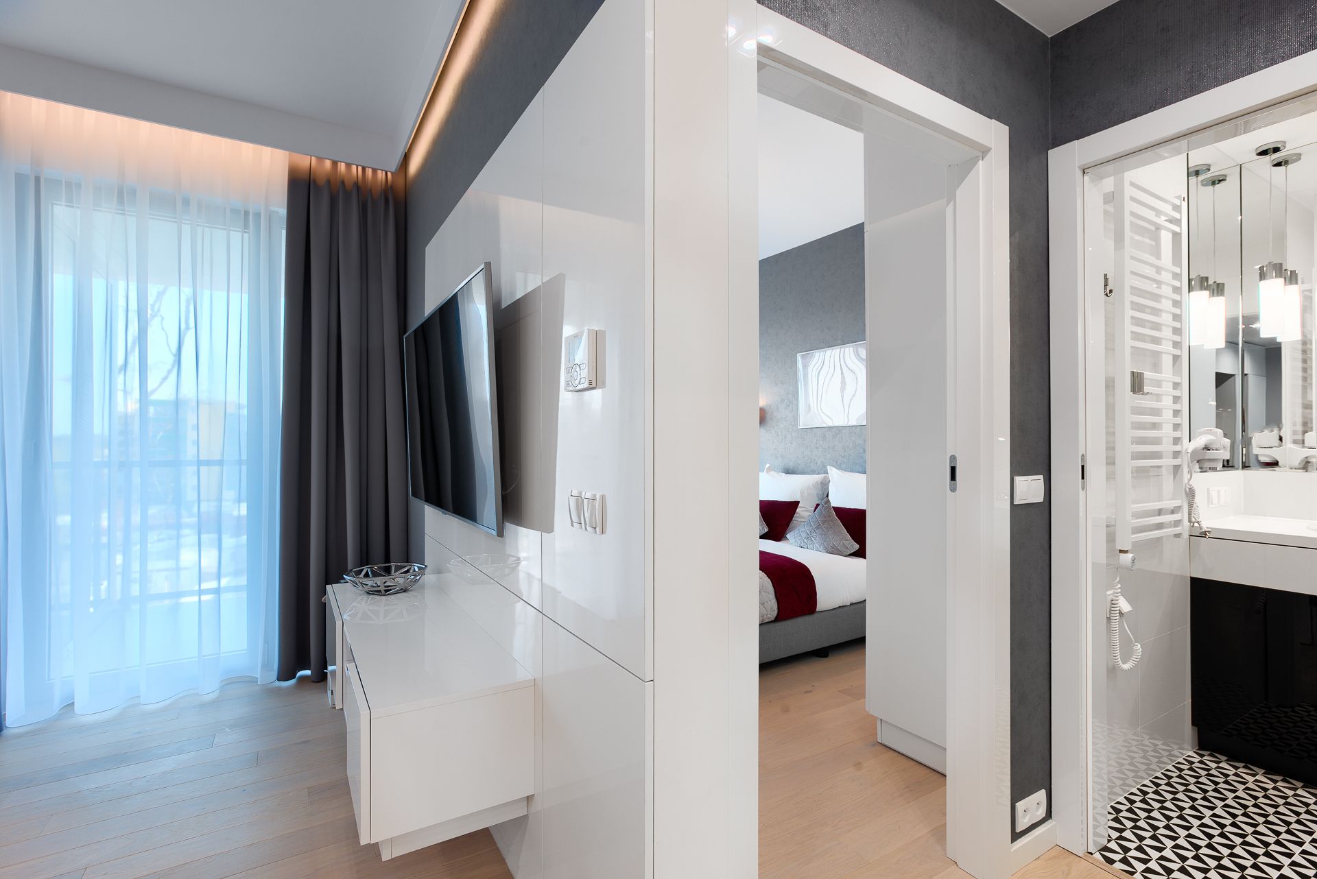 Una habitación de hotel moderna con una gran televisión de pantalla plana, una mesita de noche, una cama con ropa de cama roja y blanca, un vestidor y un baño con espejo y lavamanos.
