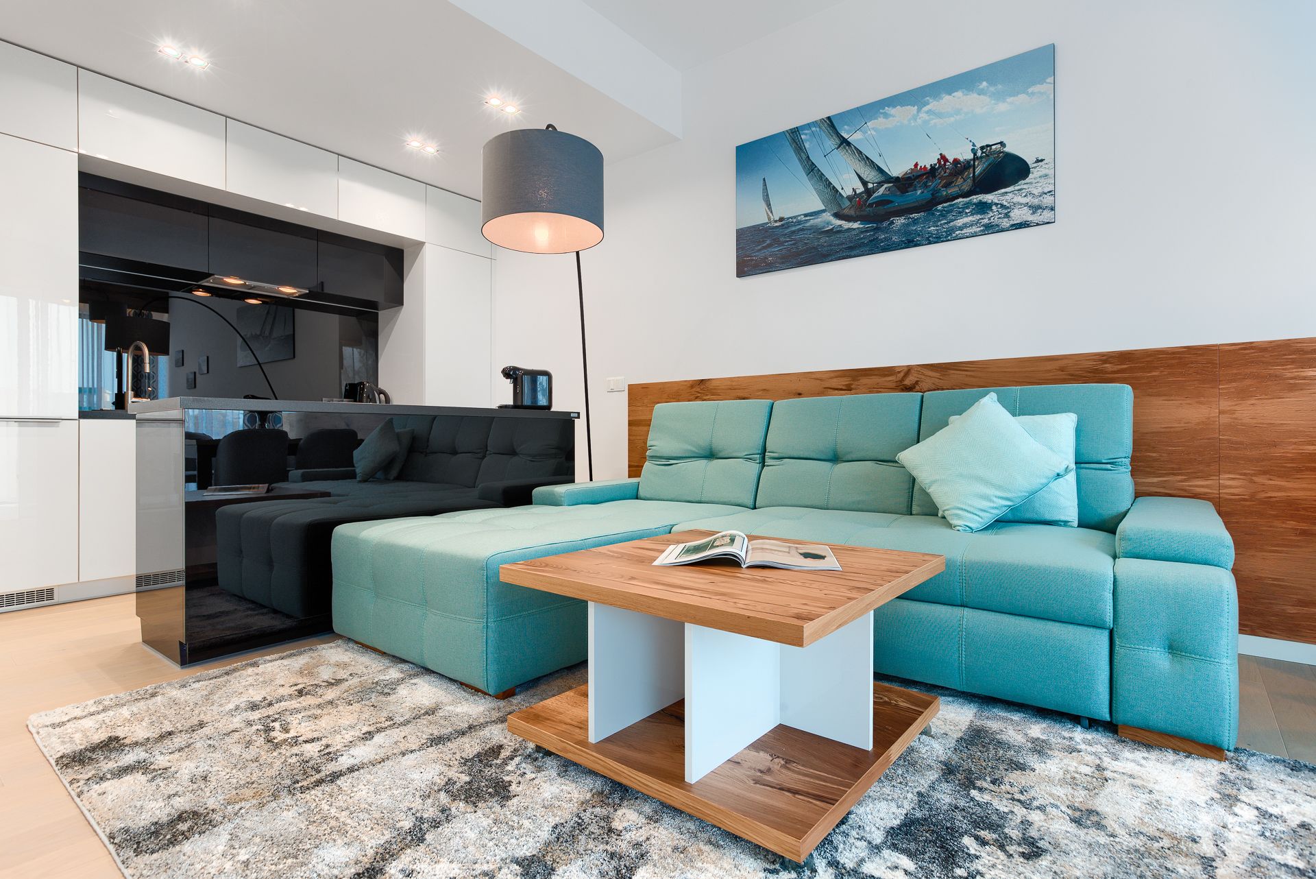 A modern living room with a teal sofa, a wooden coffee table, and a kitchen area with white cabinets and a black range hood.