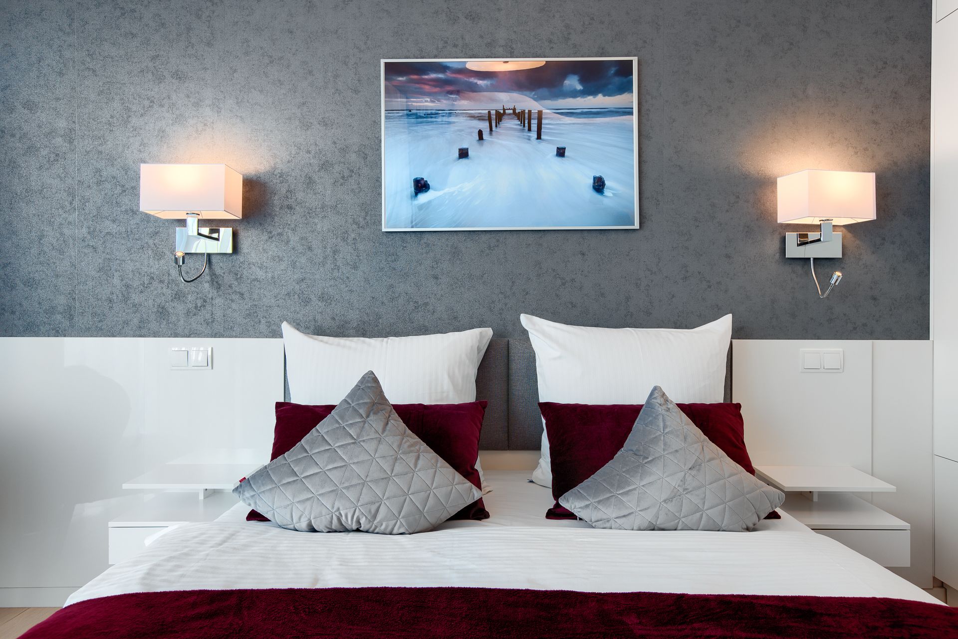 Una habitación de hotel moderna con una cama grande, dos mesitas de noche, una pared gris con textura y un cuadro enmarcado de un paisaje nevado.