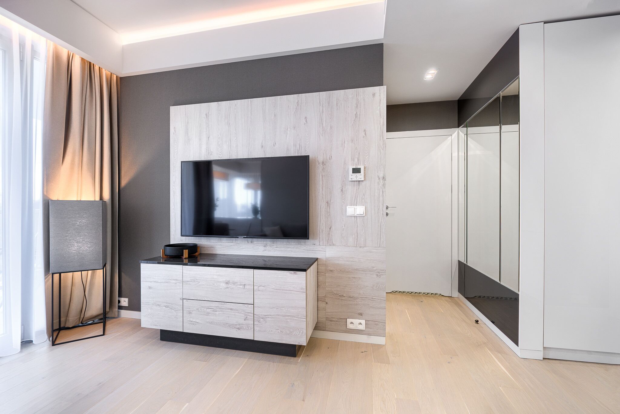 A modern living room with a flat-screen TV mounted on a light wood panel wall, a white cabinet with drawers, and a large wardrobe with mirrored doors.