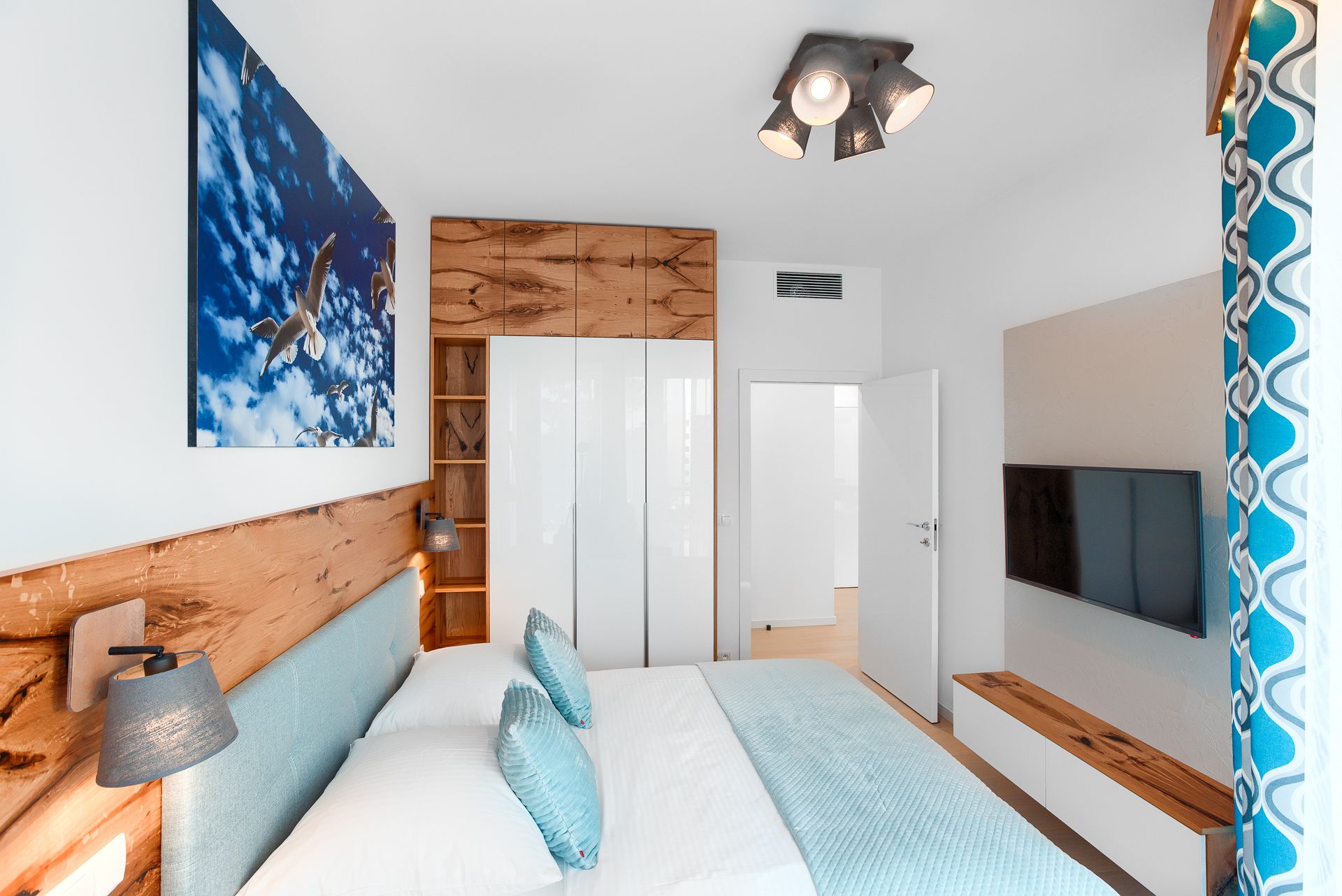 Une chambre moderne avec un lit, une tête de lit en bois, une lampe fixée au mur, un luminaire au plafond, une télévision et une armoire.