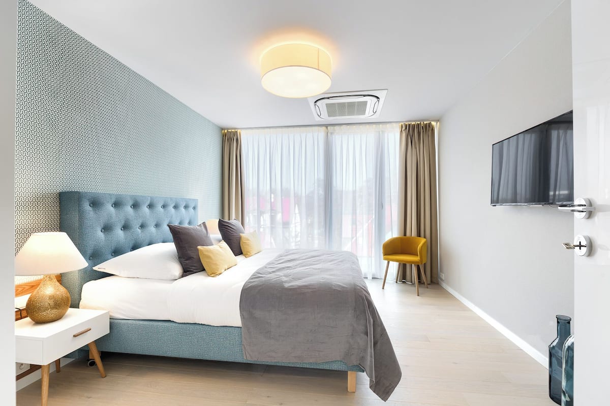 Une chambre d’hôtel moderne avec une tête de lit capitonnée, un grand lit, une table de chevet avec une lampe, une chaise et un téléviseur fixé au mur.