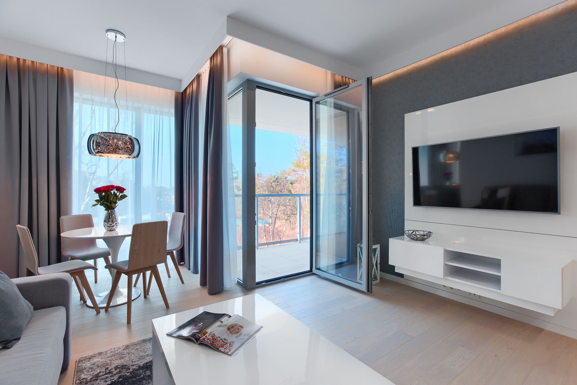 A modern living room with a large TV, a dining area with a table and chairs, and a view through a glass door.
