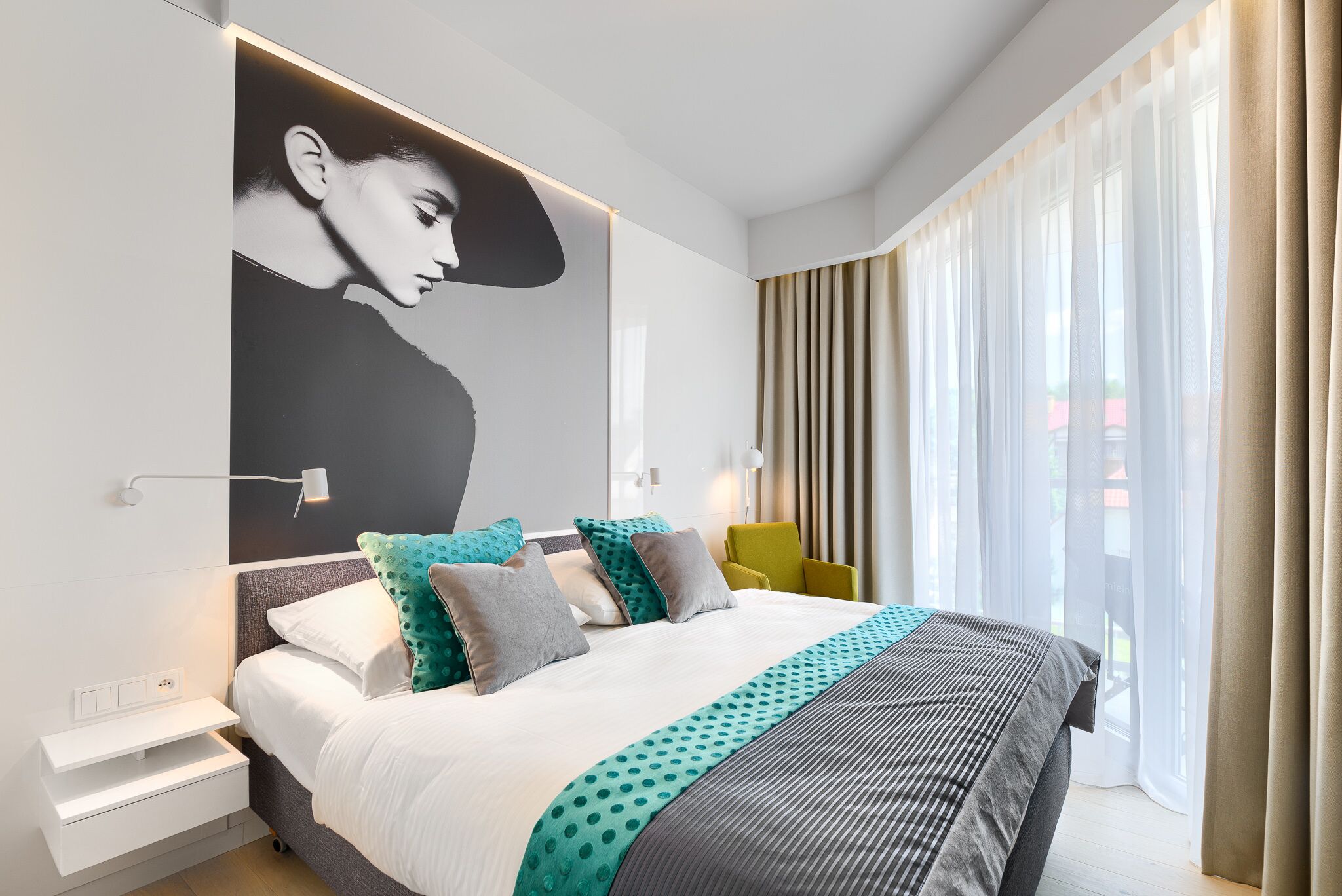 A modern hotel room with a large bed, a nightstand, a wall-mounted lamp, and a window with curtains.