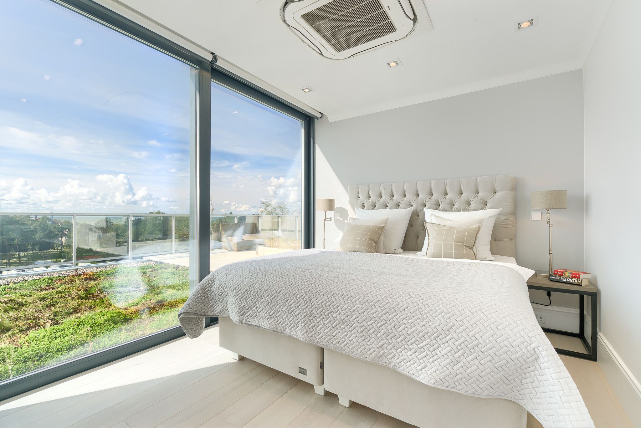A modern bedroom with a large bed, a view of greenery through floor-to-ceiling windows, and a cityscape in the distance.
