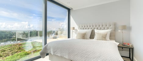 A modern bedroom with a large bed, a view of greenery through floor-to-ceiling windows, and a cityscape in the distance.
