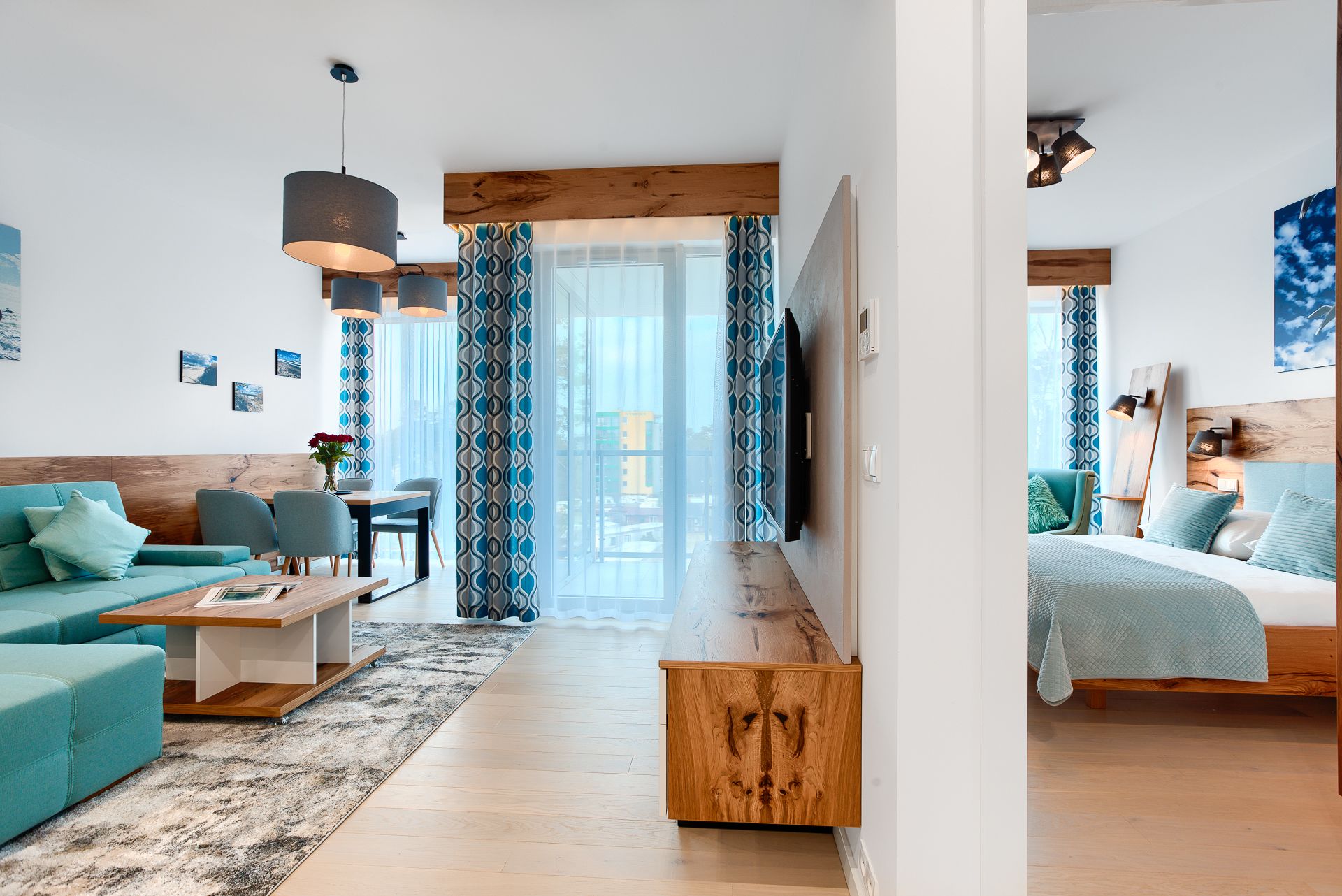 Una sala de estar moderna con una mesa de centro de madera, un sofá color turquesa y una zona de comedor con sillas. Un televisor está montado en la pared y hay un gran ventanal con cortinas estampadas.