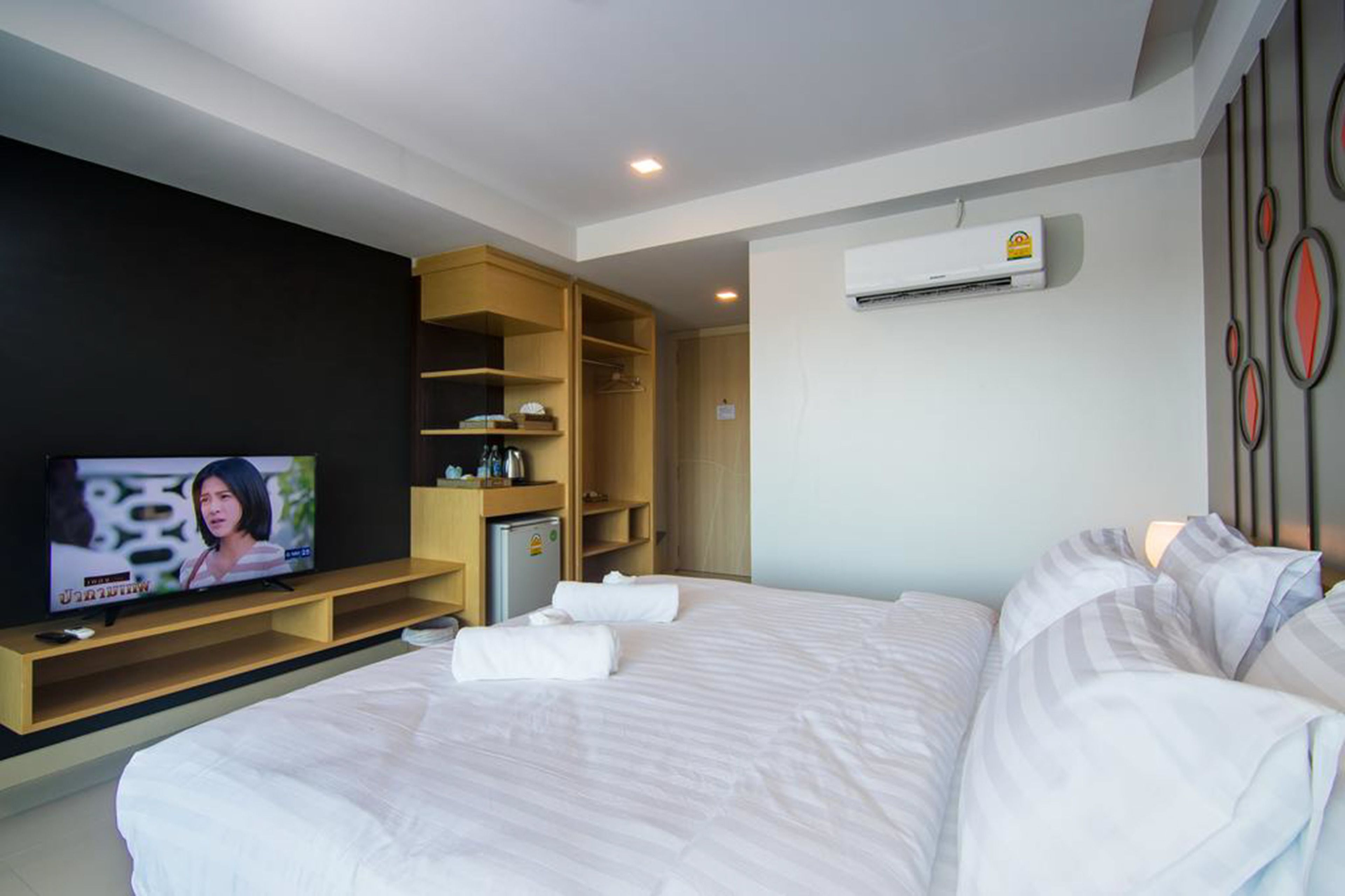 A hotel room with a large bed, a TV on a wooden stand, a mini fridge, and an air conditioning unit on the wall.