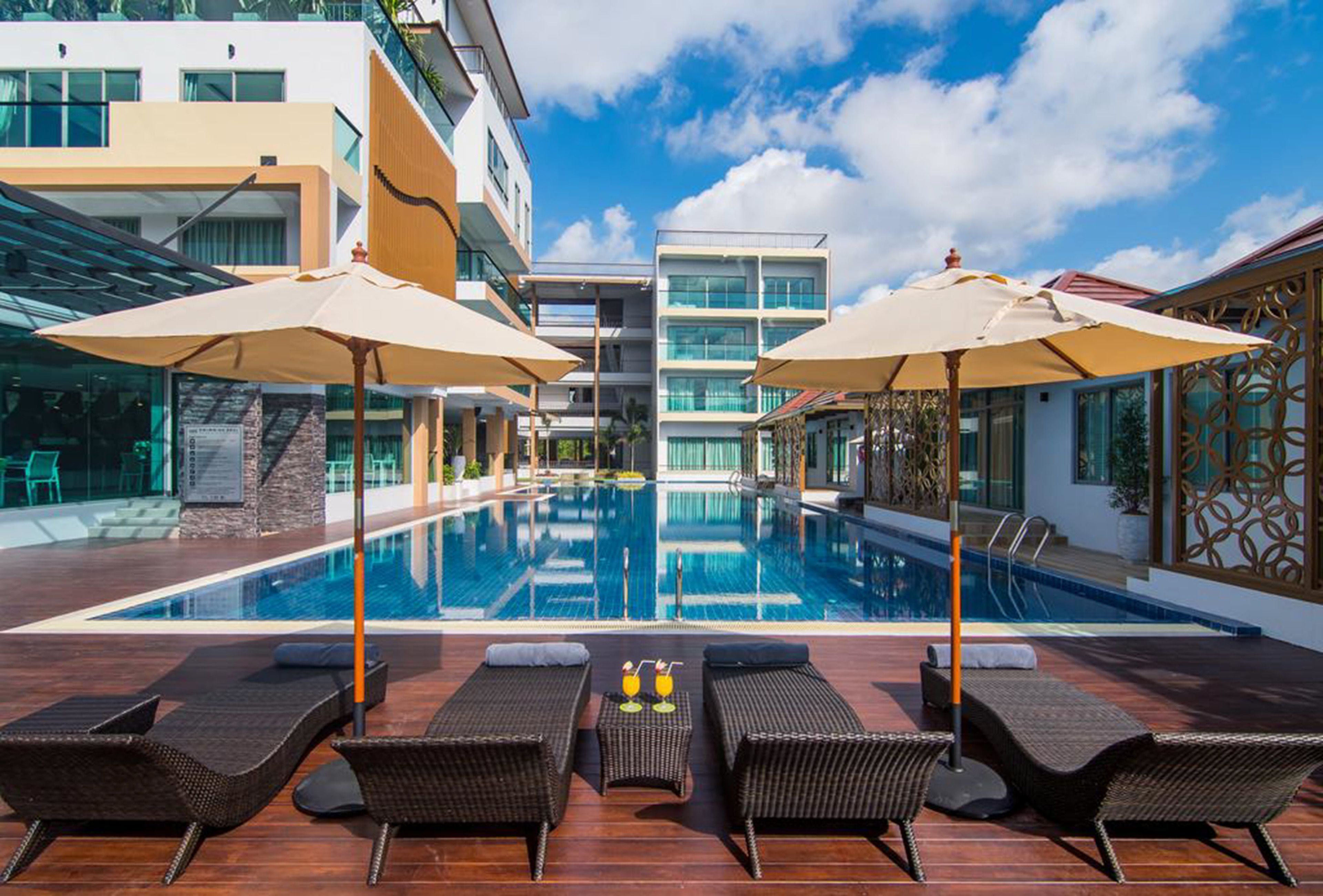 Une piscine d’hôtel moderne, dotée de chaises longues et de parasols, avec un ciel bleu azur et son reflet dans l’eau.