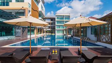Une piscine d’hôtel moderne, dotée de chaises longues et de parasols, avec un ciel bleu azur et son reflet dans l’eau.