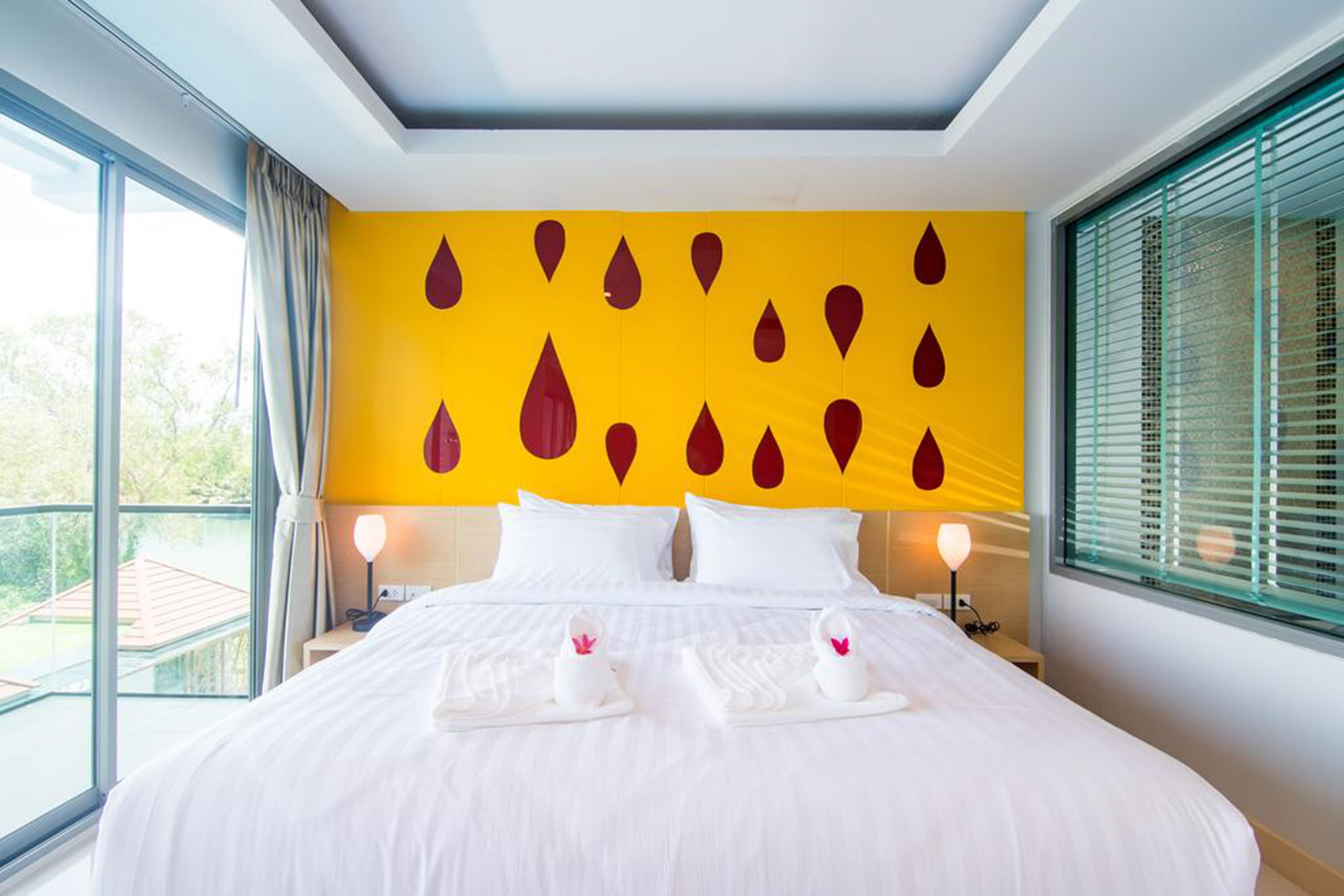 A hotel room with a large bed, two bedside tables with lamps, a yellow wall with red teardrop patterns, and a balcony view.