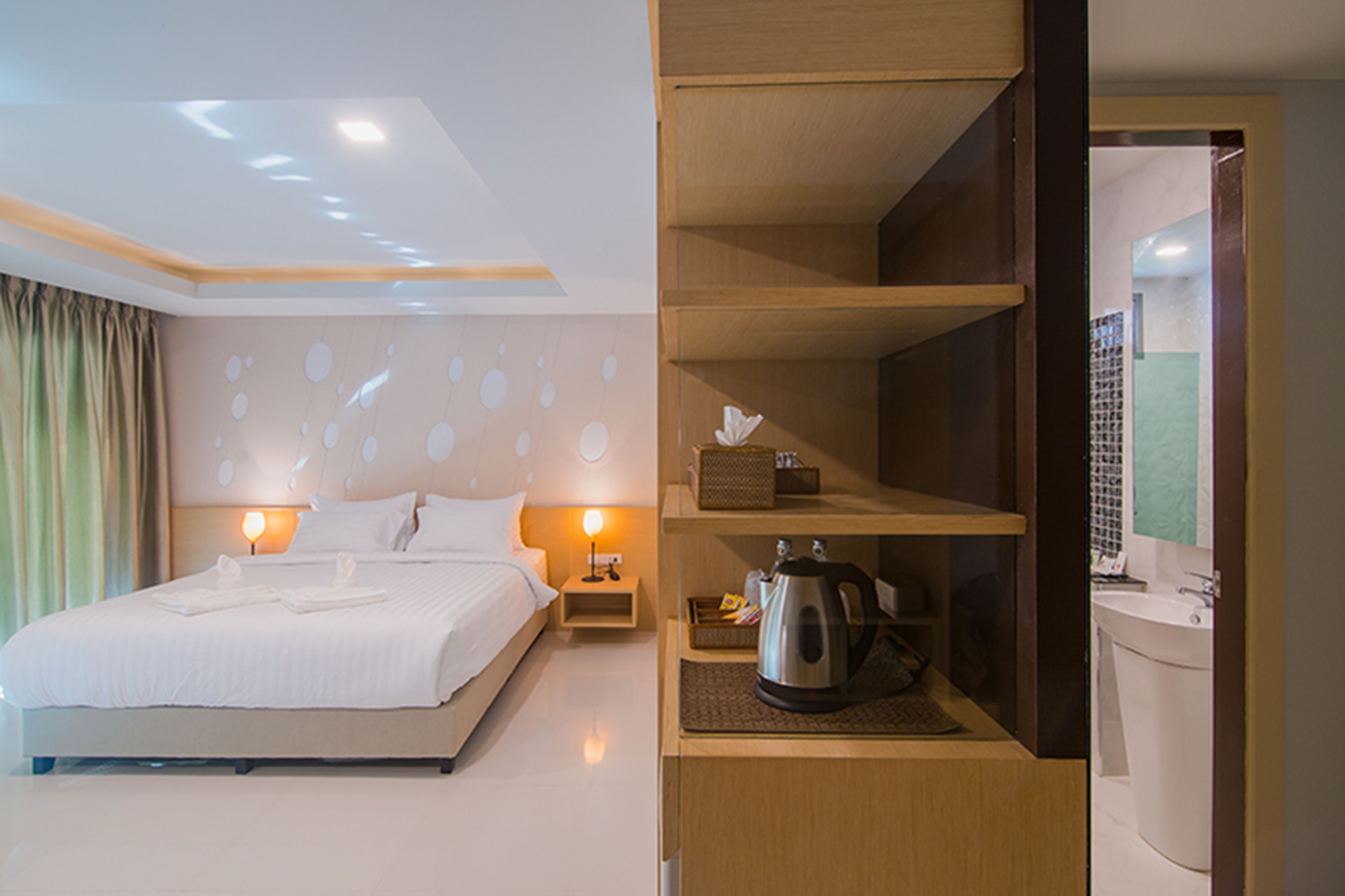 Una habitación de hotel con una cama bien hecha, una mesita de noche con una lámpara, una estantería con una tetera y una caja, y un baño con un lavabo y un espejo.