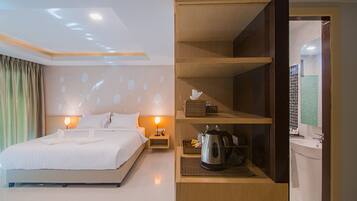 Una habitación de hotel con una cama bien hecha, una mesita de noche con una lámpara, una estantería con una tetera y una caja, y un baño con un lavabo y un espejo.