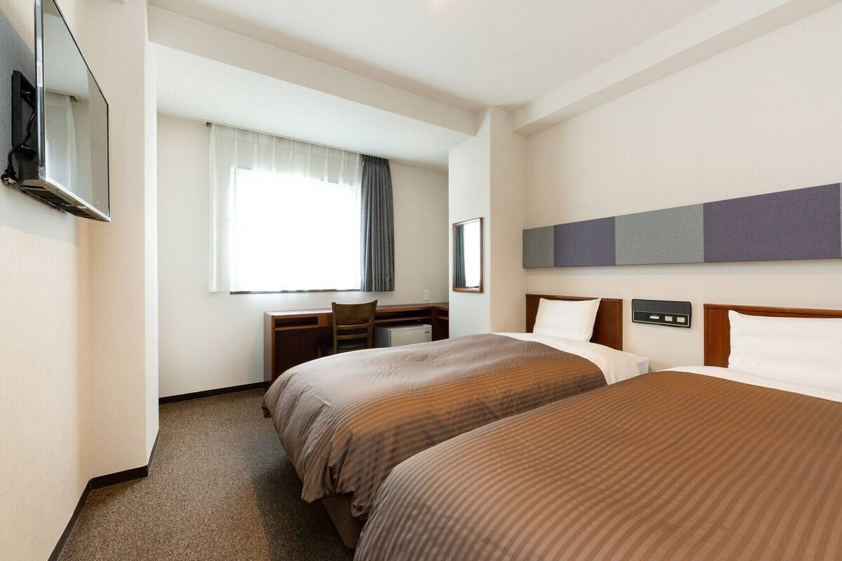 A hotel room with two beds, a desk, a chair, a TV, and a window with curtains.